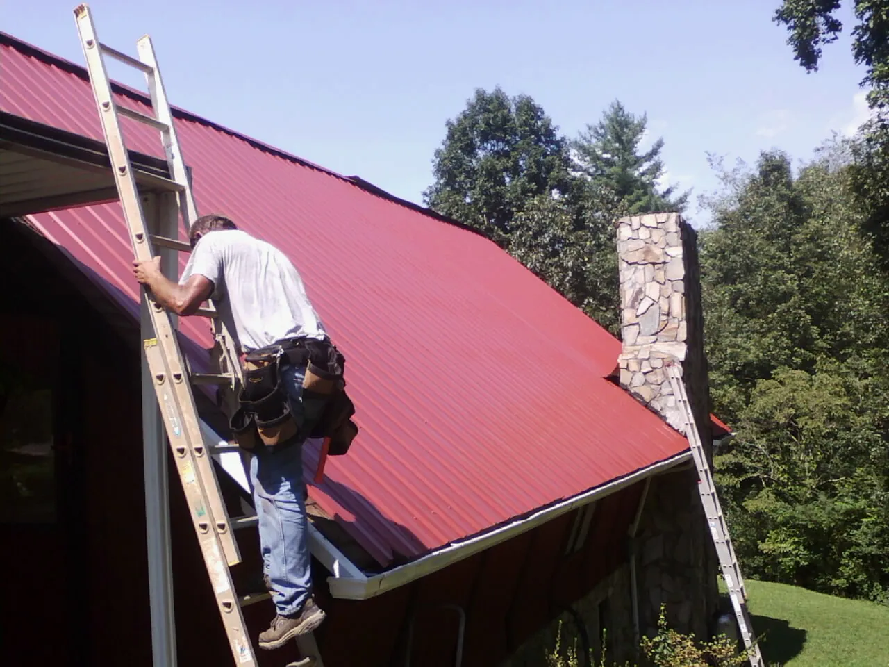 Roofer on a ladder beside a red metal roof and stone chimney — Soffit & Fascia Repair by Roofing Brothers in Hoosick Falls